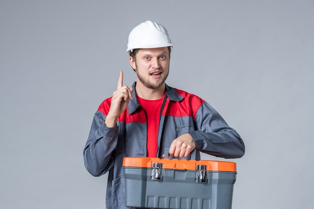 front view male builder in uniform and helmet holding tool case on gray background