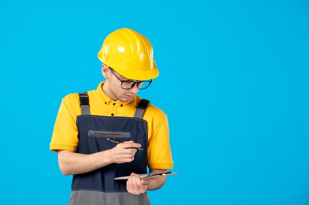 Vista frontale del costruttore maschio in uniforme e casco che prende appunti sulla parete blu