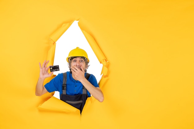 Front view male builder in uniform holding black bank card on yellow background