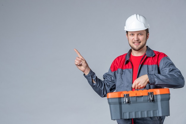 front view male builder in uniform holding heavy tool case on gray background
