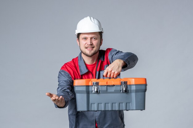 мужчина-строитель в униформе, держащий чемодан с инструментами на сером фоне, вид спереди
