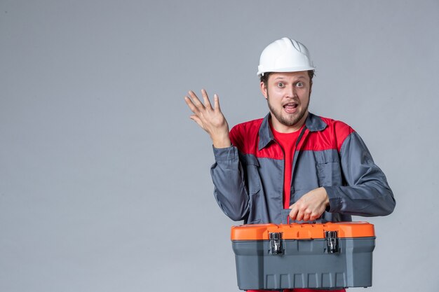 мужчина-строитель в униформе, держащий чемодан с инструментами на сером фоне, вид спереди