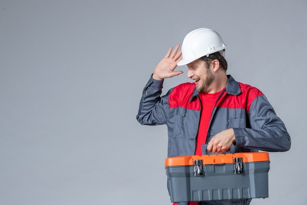 Vista frontale costruttore maschio in uniforme che tiene la valigetta degli attrezzi e ride su sfondo grigio