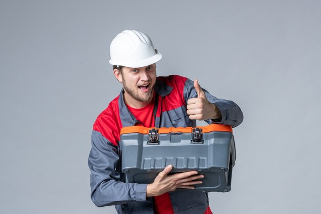 front view male builder in uniform trying to open tool case on gray background