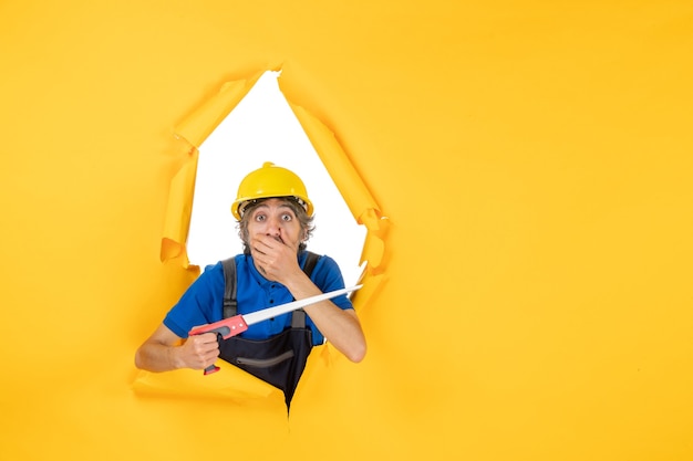 Front view male builder in uniform with instrument in his hands shocked on yellow wall job building worker constructor color work