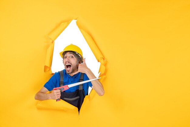 黄色の壁の建物の労働者の仕事のコンストラクターの色に彼の手で楽器と制服を着た正面図の男性ビルダー