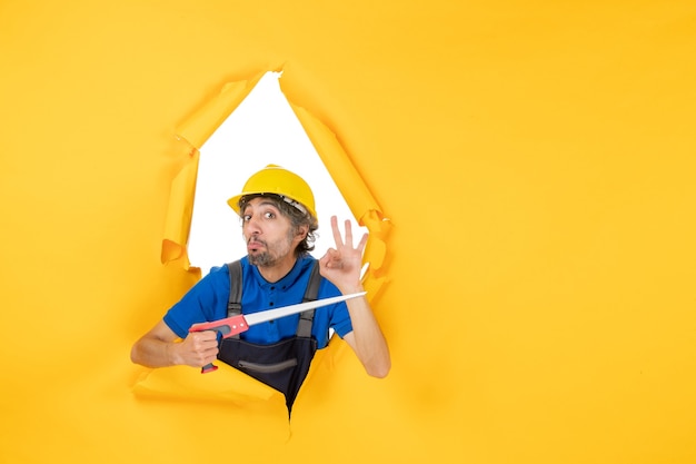 Front view male builder in uniform with instrument in his hands on yellow wall building worker job constructor work