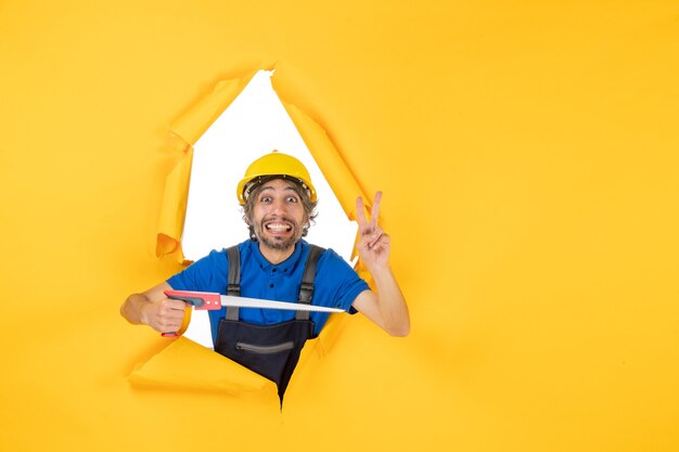 Costruttore maschio di vista frontale in uniforme con lo strumento nelle sue mani su un lavoratore di costruzione di colore di lavoro del costruttore di lavoro della parete gialla