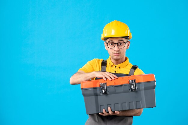 Front view of male builder in uniform with tool box in his hands on blue surface