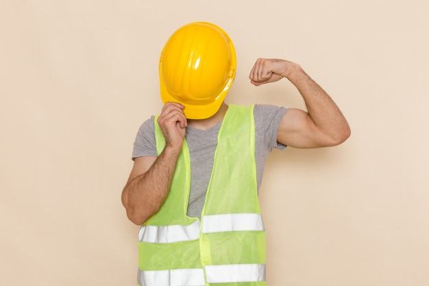 Front view male builder in yellow helmet hiding his face flexing on light desk