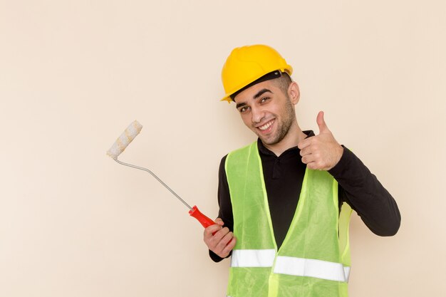 Вид спереди мужчина-строитель в желтом шлеме, держащий кисть для стен, улыбаясь на светлом фоне