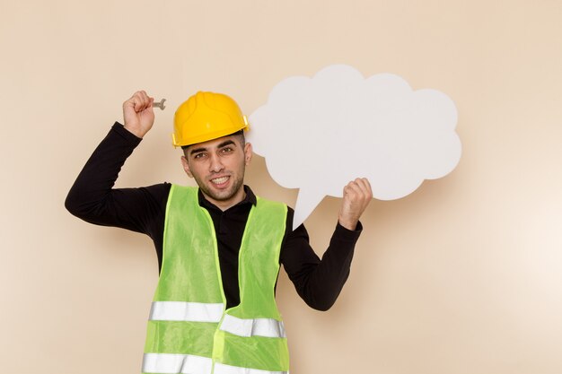 Front view male builder in yellow helmet holding tool and white sign on cream background