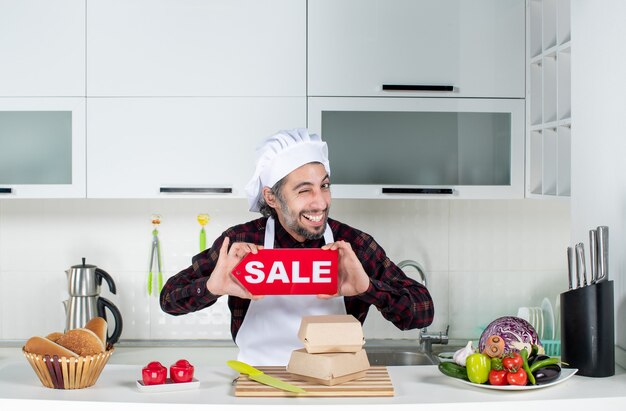 Front view of male chef blinking eye holding up sale sign in the kitchen