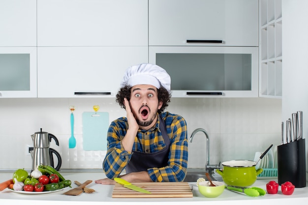 Vista frontale dello chef maschio che cucina verdure fresche sentendosi scioccato nella cucina bianca