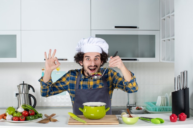 Front view of male chef cooking fresh vegetables tasting ready meal and making eyeglases gesture in the white kitchen