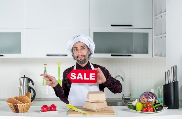 Front view of male chef pointing at left holding up sale sign in the kitchen