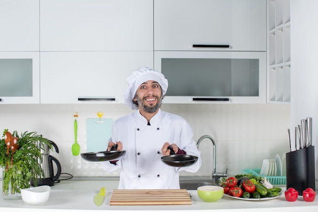 Front view male chef in uniform holding up different size pans in modern kitchen