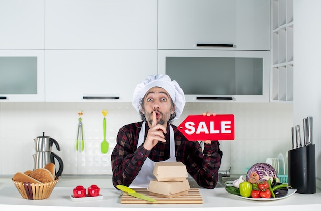 Vista frontale dello chef maschio in uniforme che regge il cartello rosso di vendita che fa segno di silenzio nella cucina moderna