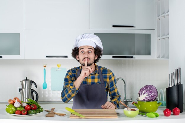 新鮮な野菜とキッチンツールで調理し、白いキッチンで沈黙のジェスチャーを作る男性シェフの正面図