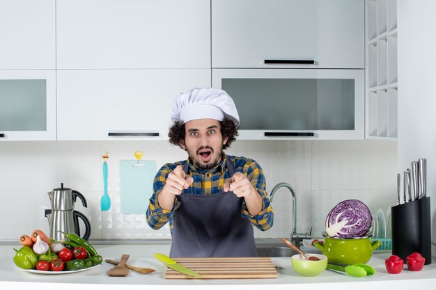Вид спереди шеф-повара-мужчины со свежими овощами и готовкой с кухонными принадлежностями и указывая вперед на белой кухне