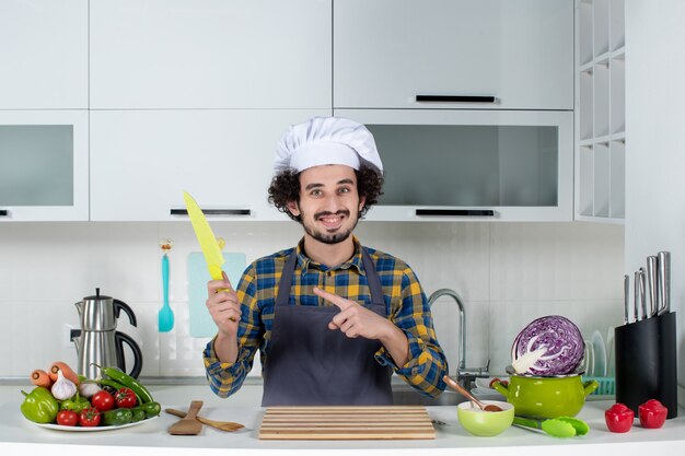 Front view of male chef with fresh vegetables and cooking with kitchen tools and pointing up holding knife in the white kitchen