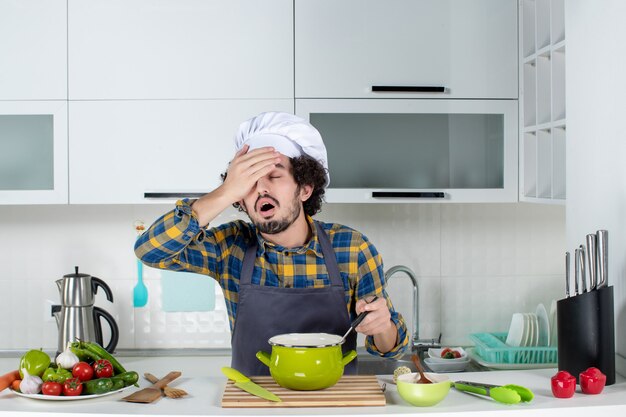 新鮮な野菜と白いキッチンで疲れた感じの食事でスプーンを保持している男性シェフの正面図