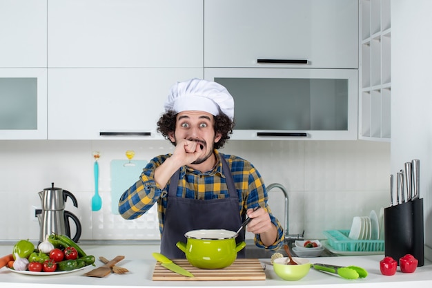 Vista frontale dello chef maschio con verdure fresche e tenendo il cucchiaio nel pasto sentendosi spaventato nella cucina bianca