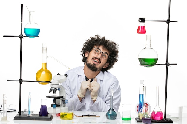 Вид спереди мужской химик в медицинском костюме, просто сидящий перед столом с растворами на светлом белом пространстве