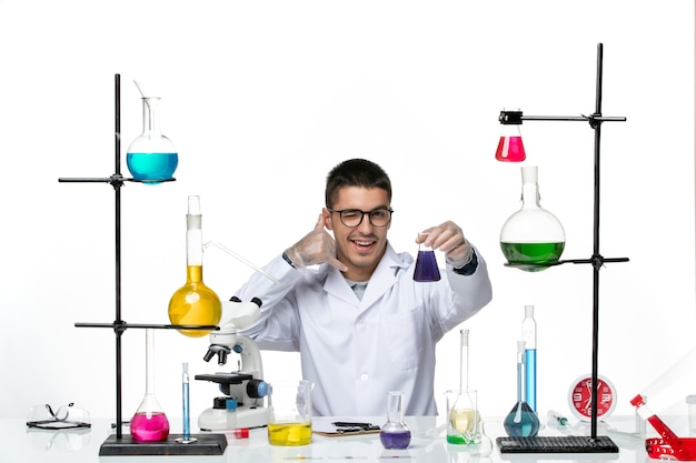 Front view male chemist in medical suit sitting and holding flask with solution on light-white background virus covid splash disease science