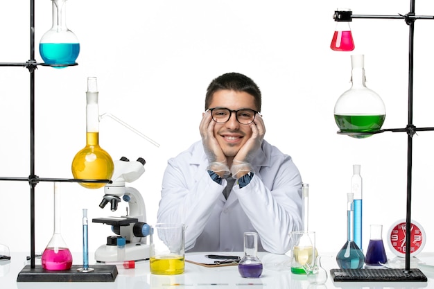 Front view male chemist in medical suit sitting and smiling on white background virus covid splash disease science