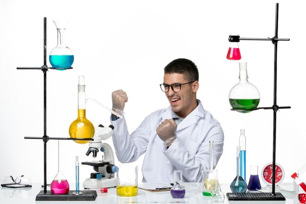 Front view male chemist in medical suit sitting with solutions and rejoicing on white background virus covid disease science