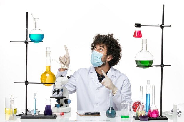 Вид спереди мужской химик в медицинском костюме и с маской, просто сидящий с разными растворами, позирует на светлом белом пространстве