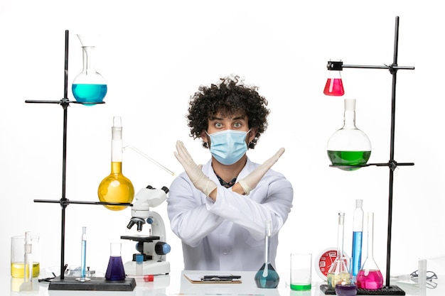 Front view male chemist in medical suit and with mask just sitting with different solutions posing on a white space