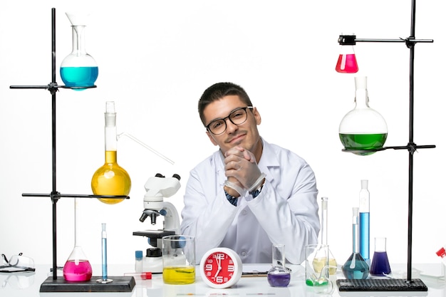 Вид спереди мужской химик в белом медицинском костюме сидит и улыбается на белом фоне вирусная наука covid- пандемическая лаборатория