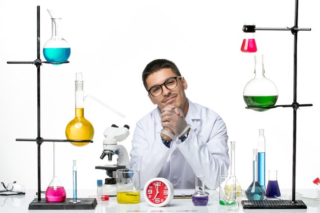 Front view male chemist in white medical suit sitting and smiling on white background virus science covid- pandemic lab