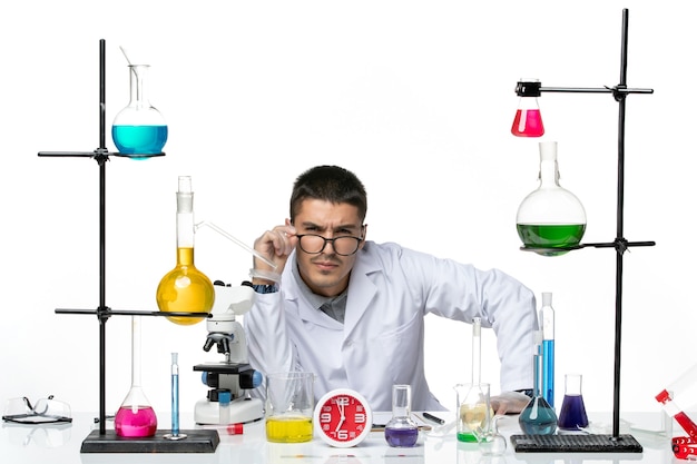 Вид спереди мужской химик в белом медицинском костюме, сидящий с разными растворами на белом фоне, научная лаборатория вирусных заболеваний covid