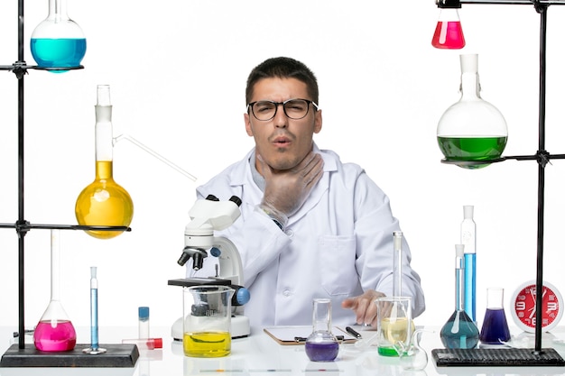 Вид спереди мужской химик в белом медицинском костюме, сидящий с растворами, имеющими боль в горле, на белом фоне вирусная лаборатория covid- болезнь наука