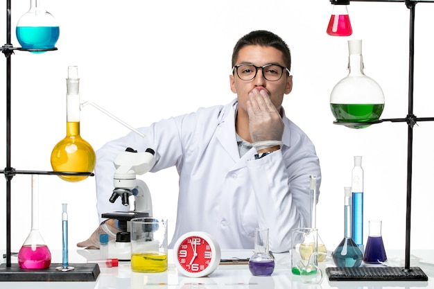 Вид спереди мужской химик в белом медицинском костюме, сидящий с растворами на белом фоне, лаборатория вирусной науки, пандемия covid