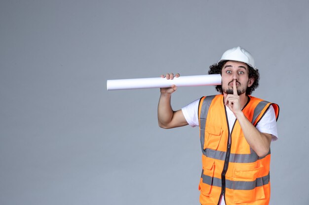 Front view of male constructor in warning vest wearing safety helmet and holding blank making silence gesture on gray wave wall