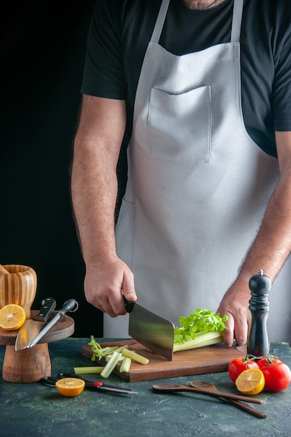 Вид спереди мужской повар, режущий сельдерей на темной стене, салат, здоровая диета, цветная фотография