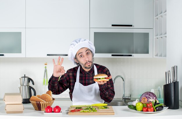 Вид спереди мужского повара, жестикулирующего, подняв гамбургер, стоящего за кухонным столом