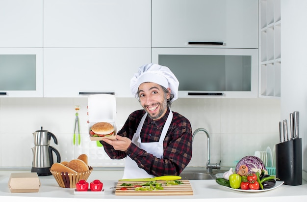 Front view of male cook holding up burger standing behind kitchen table in modern kitchen