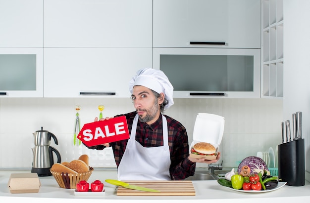 Front view of male cook holding up sale sign and burger in the kitchen