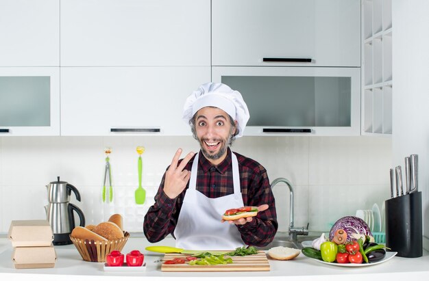 Front view of male cook making burger showing v gesture standing behind kitchen table