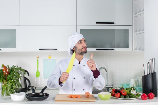 Мужчина-повар, вид спереди в униформе, держит нож, удивляет своей идеей на кухне