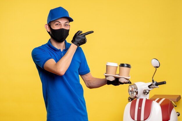 黄色の配達の仕事の共同パンデミックサービスの制服でコーヒーを保持している黒いマスクの正面図の男性の宅配便