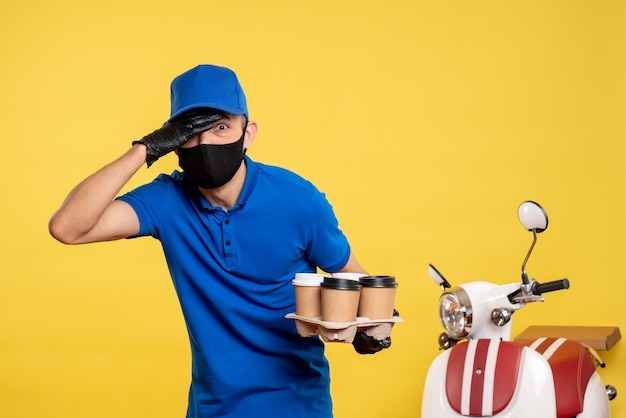 Курьер-мужчина в черной маске, держащий кофе на желтой работе, вид спереди - работа по доставке пандемических служб