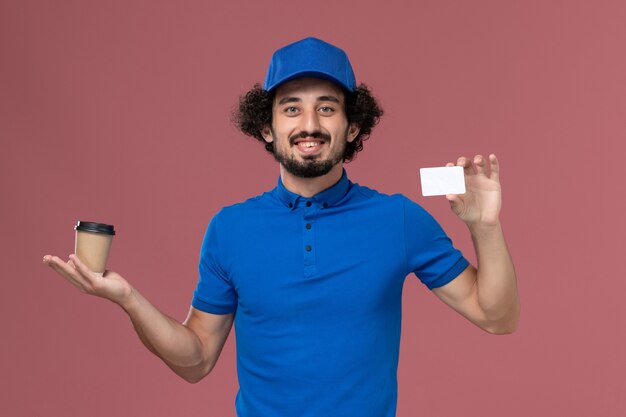 Front view of male courier in blue uniform and cap with delivery coffee cup and card on his hands on the pink wall