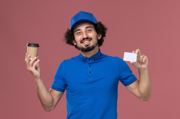 Front view of male courier in blue uniform and cap with delivery coffee cup and card on his hands on the pink wall