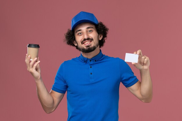 青い制服とピンクの壁に彼の手に配達コーヒーカップとカードとキャップの男性宅配便の正面図